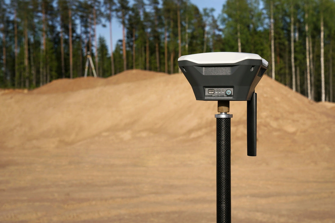 A GNSS antenna attached to a GNSS receiver standing on a pole with a sandy background and palm threes behind.