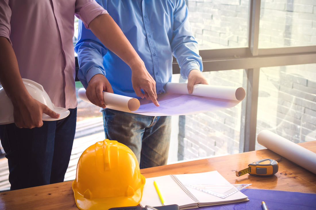 Two individuals examining architectural plans on a desk with a hard hat and measuring tools nearby, following land surveying best practices.